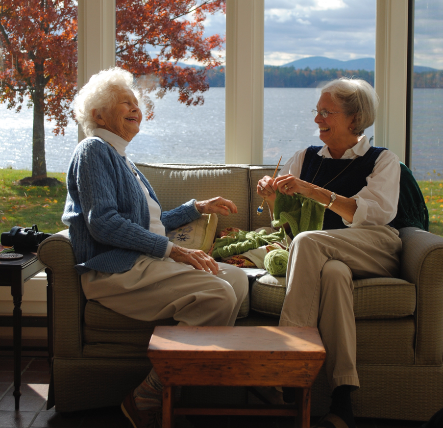 Two older smiling white women on a smal tan couch enjoying each other's company. The woman on the right is knitting and has her legs crossed. She has glasses and a short bob haircut and is wearing a blue and white shirt and tan pants. The woman on the left has a beautiful spray of short white hair and is somewhat older than the other woman. She is wearing a blue cardigan and tan pants. Behind them is a large picture window overlooking a sparkling lake. There is a tree on the grassy banks with leaves that have turned red for autumn. In the background across the lake there are low mountains and a partly cloudy sky.